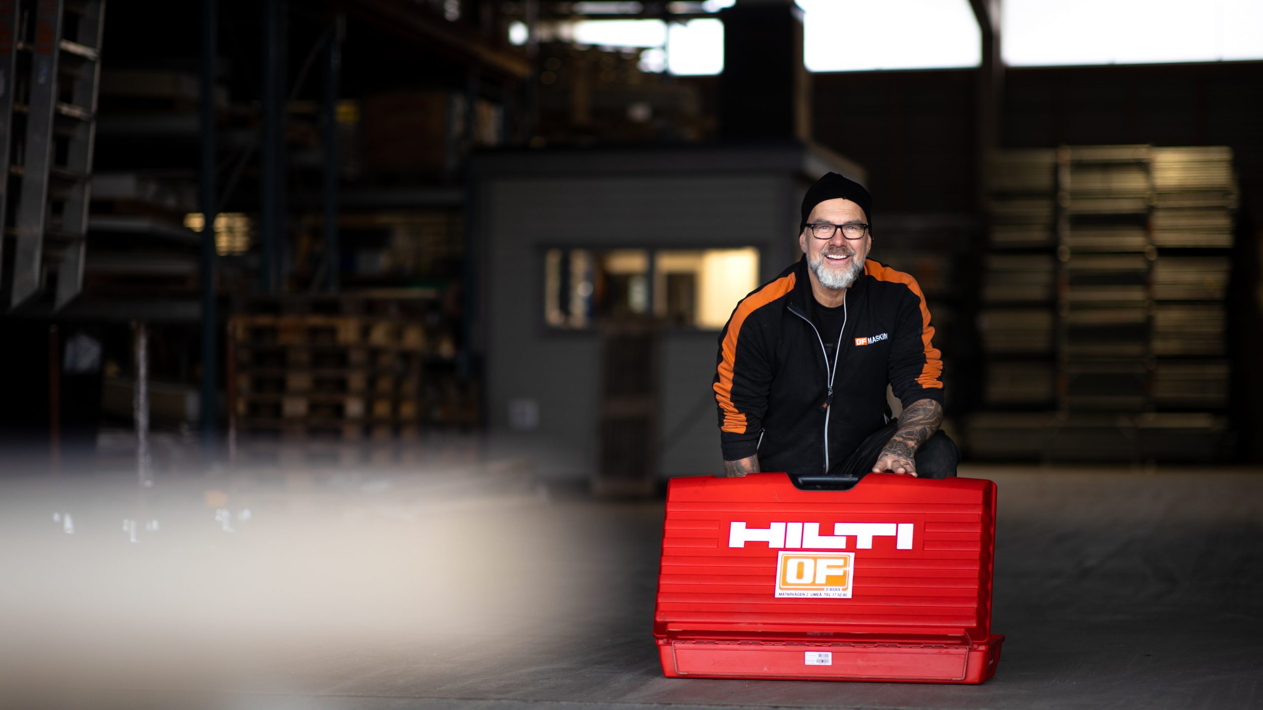 En leende man i OF Maskin-arbetskläder sitter bakom en röd Hilti-verktygslåda i en lagerlokal.
