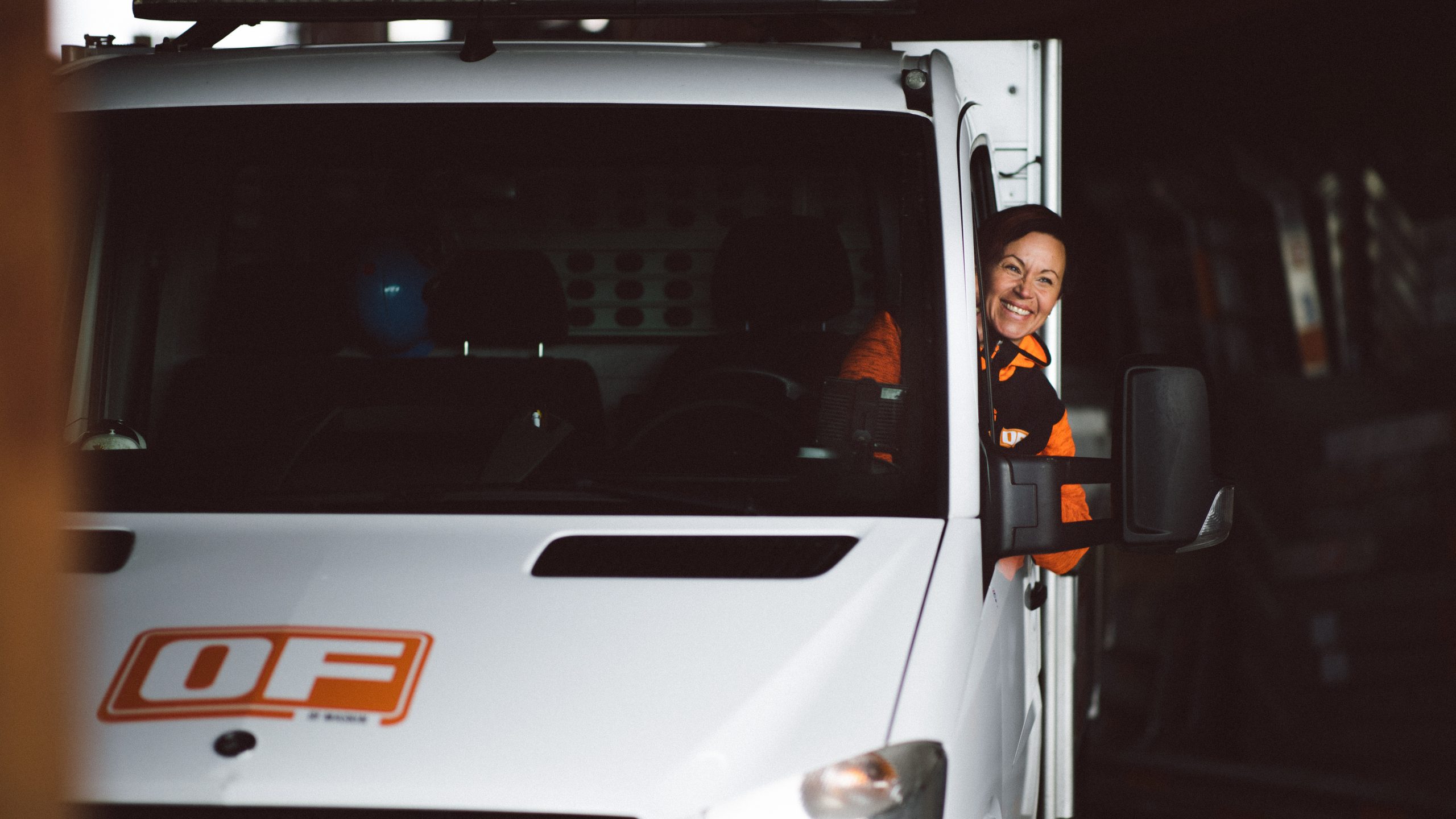 En leende förare i OF-arbetskläder tittar ut från förarsätet på en vit servicebil med OF-logotyp.