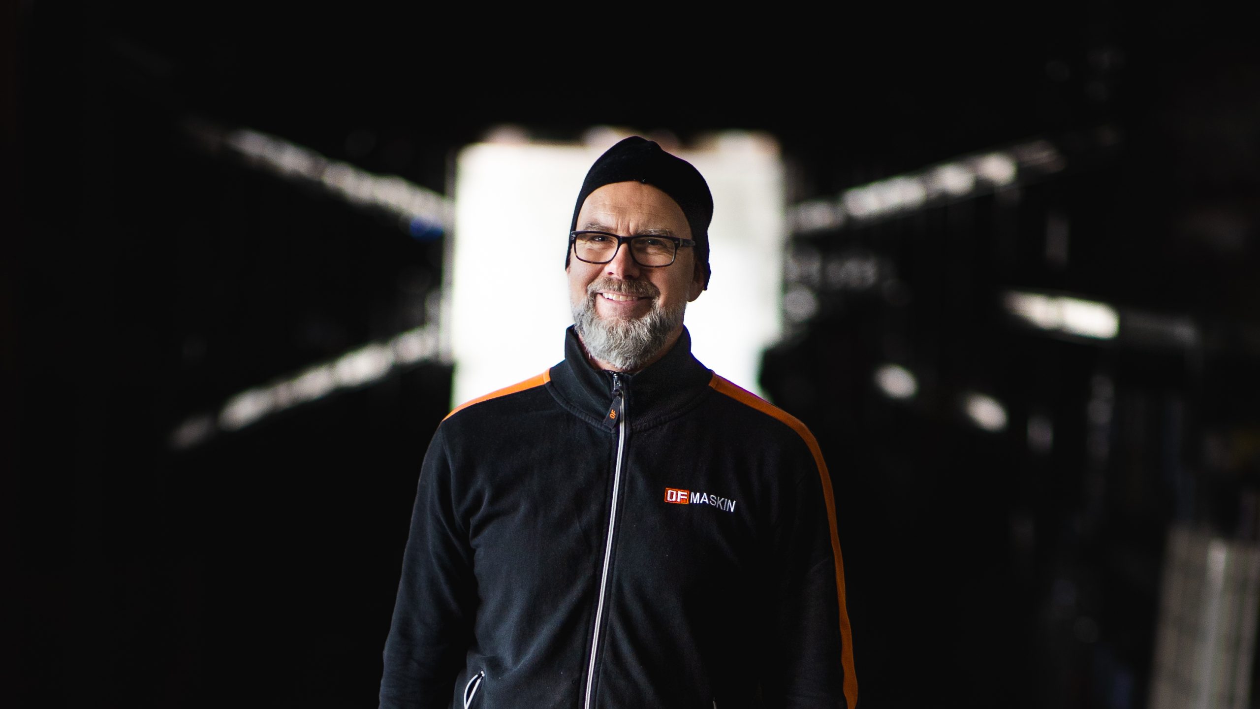 En leende arbetare på OF Maskin står mot en mörk bakgrund med ljusinsläpp.